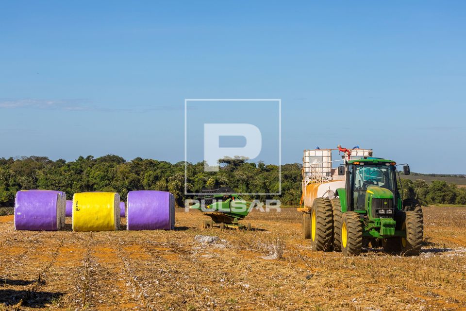 Trator puxando carreta com reservatório de água contra incêndios e à esquerda fardos de algodão recém co