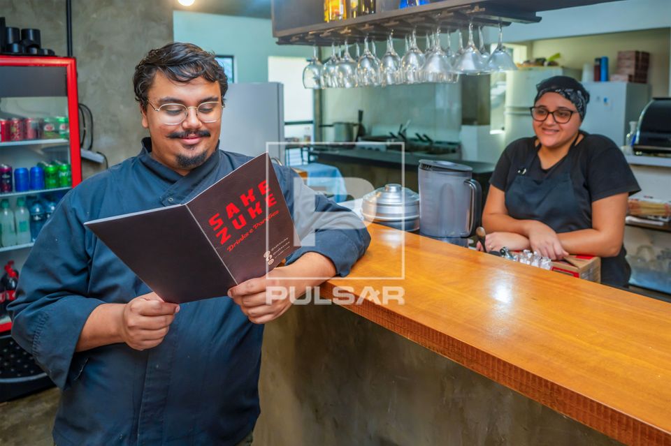 Vista interna de restaurante de comida japonesa com chef de cozinha analisando cardápio observado por ajudant