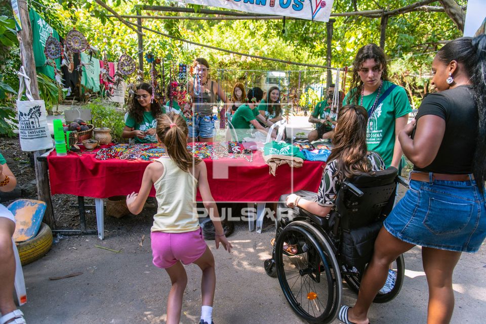 Cadeirante observando artesanato indígena à venda durante o evento Abril Indígena 2026 na aldeia urbana mul