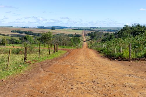 Paisagem rural com estrada de terra vermelha e área agrícola ao fundo