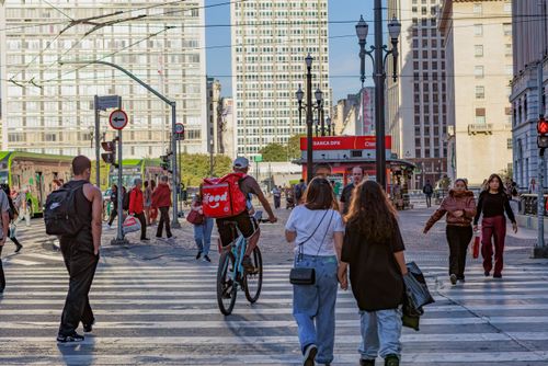 Ciclista entregador de comida por aplicativo e pessoas atravessando na Rua Xavier de Toledo com Praça Ramos d