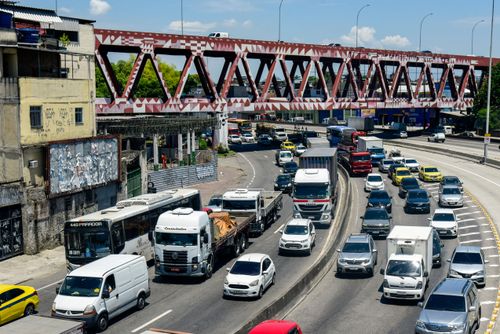 Veículos na Avenida Brasil na região do Caju–Benfica com a Linha Vermelha ao fundo e viaduto metálico com