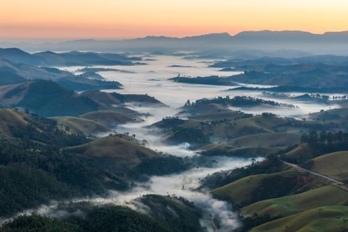 Paisagem rural com áreas de pastagem entre vales e montanhas cobertas por nevoeiro ao amanhecer na Serra da M